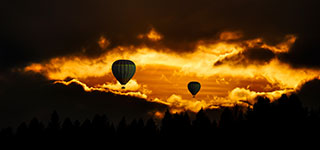 Two hot air balloons floating above trees during a vibrant orange sunset.