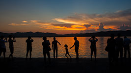Silhouettes of people standing and playing near a body of water during a vibrant orange and blue sunset.