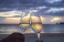 Two hands holding champagne glasses in a toast on a beach at sunset